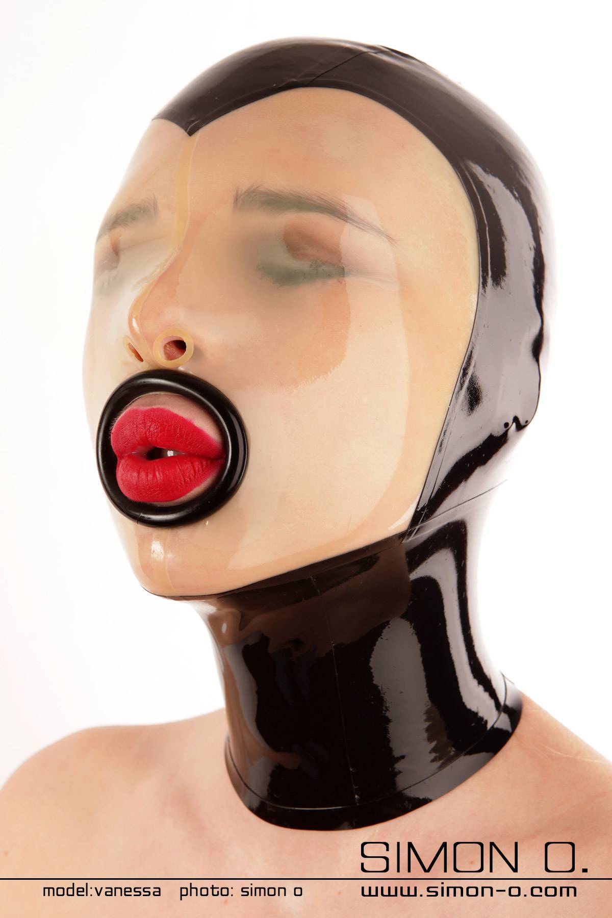 Anliegende Latex Maske in Schwarz mit transparenten Einsatz im Bereich des Gesichtes. Augen geschlossen und beim Mund befindet sich ein schwarzer Ring