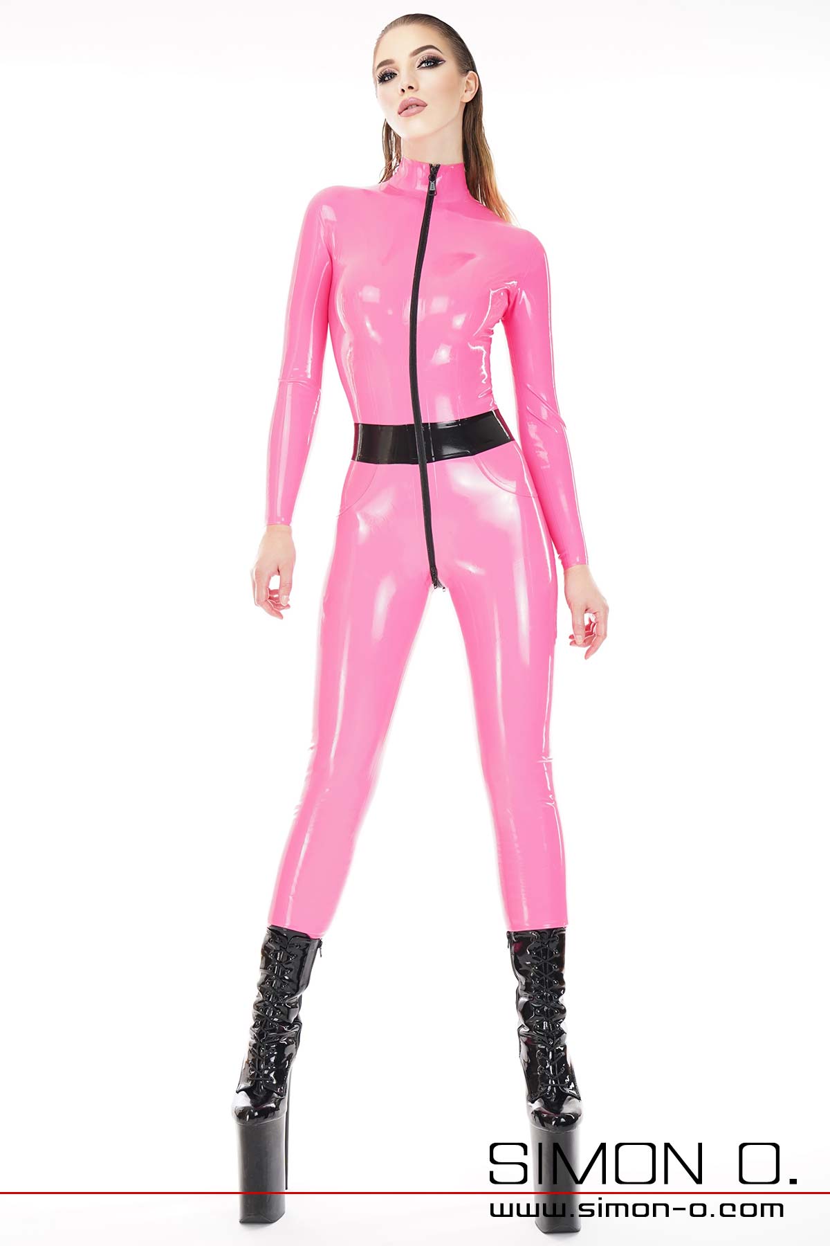 A model wears a skin-tight pink colored latex catsuit with black zipper and belt. She wears black platform high heels.