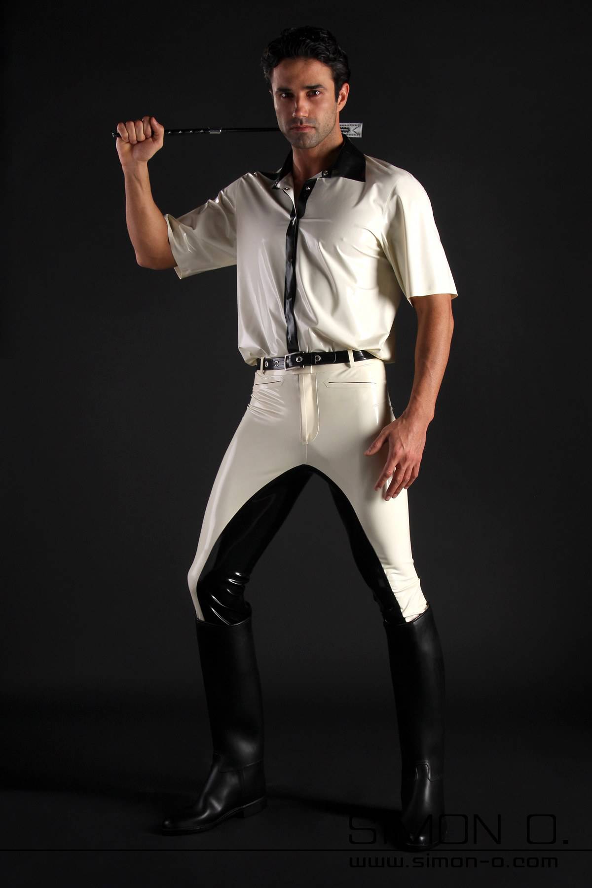 A man with a white latex shirt and a riding crop behind his head wears black and white latex breeches and riding boots.