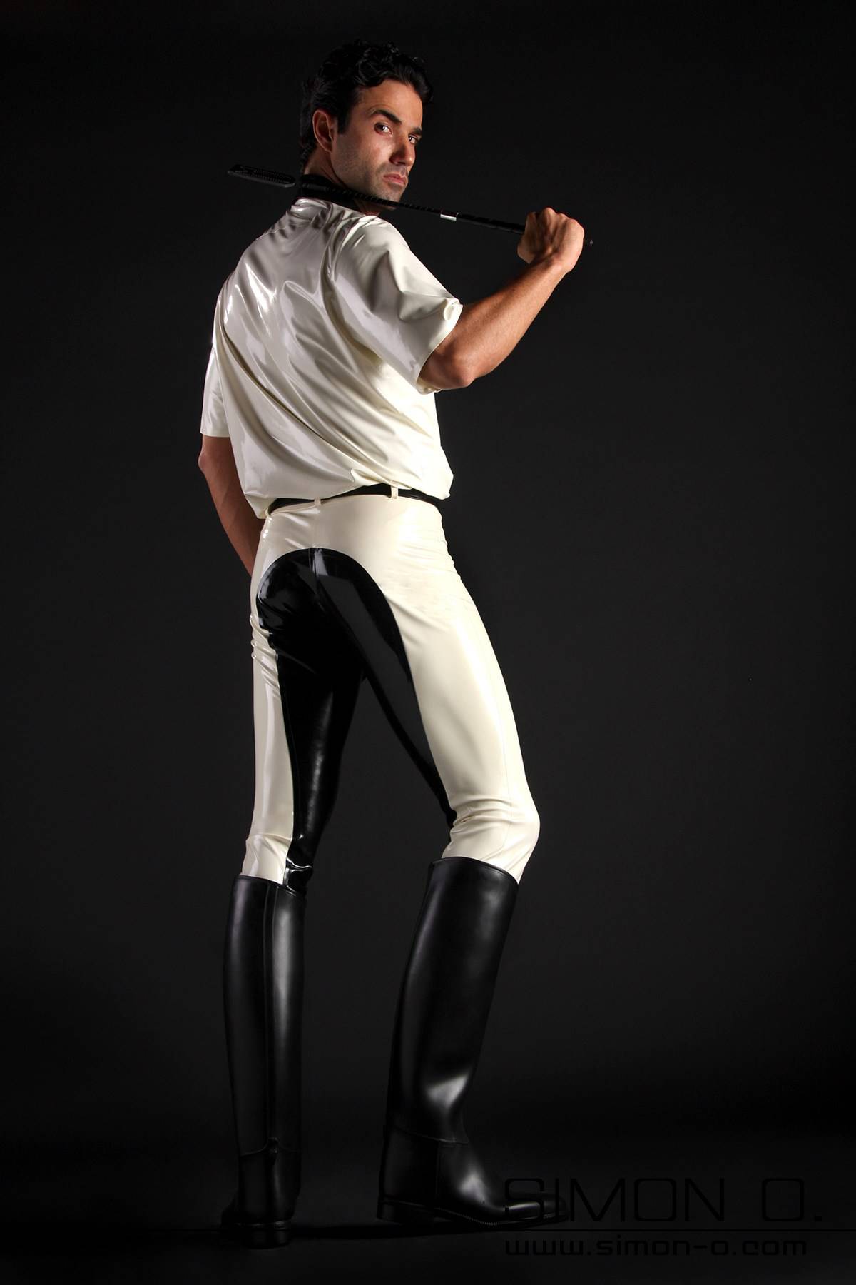 A man with a white latex shirt and a riding crop wears black and white latex breeches and riding boots - seen from behind.