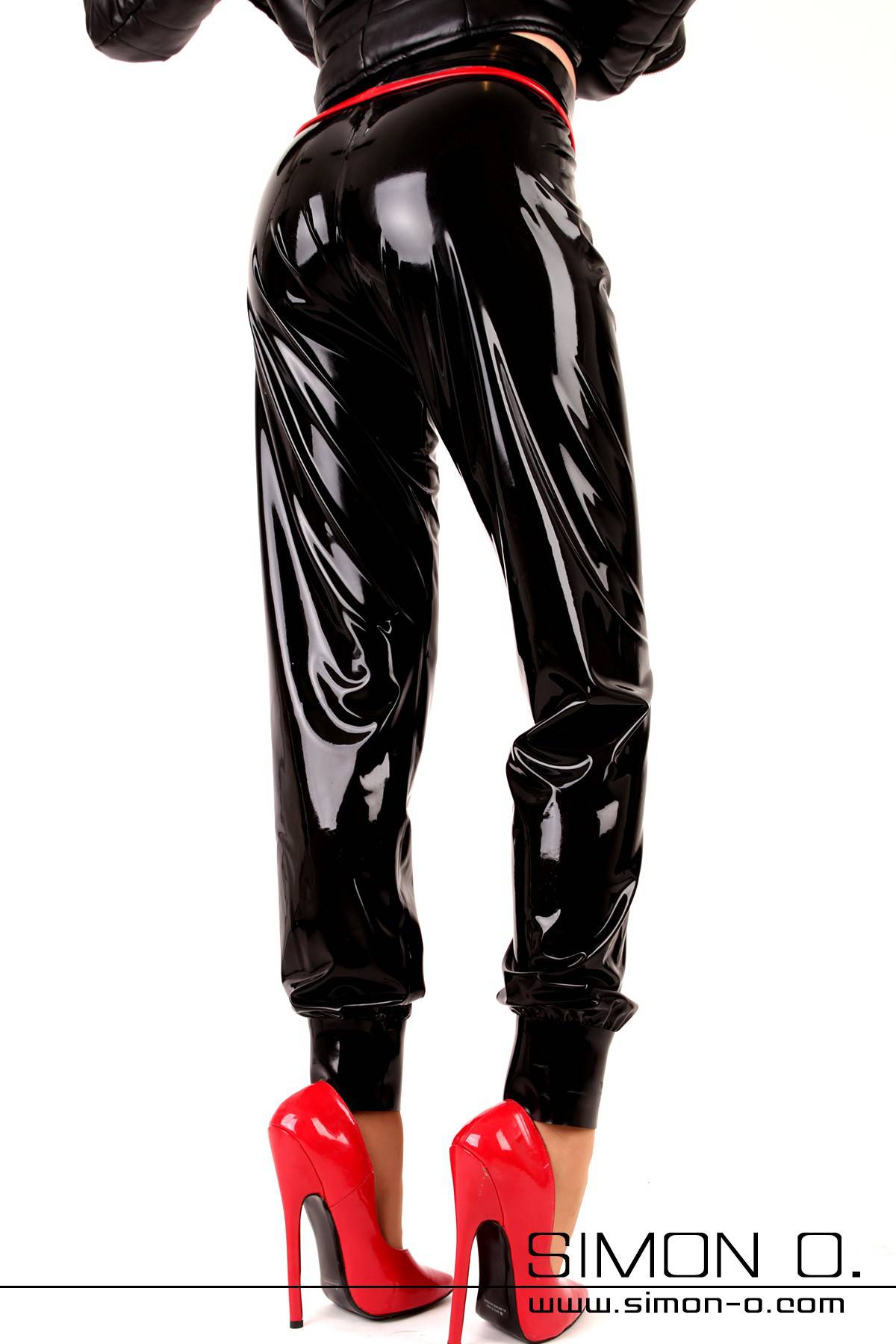 A woman wears shiny latex sweatpants in black with red high heels seen from behind