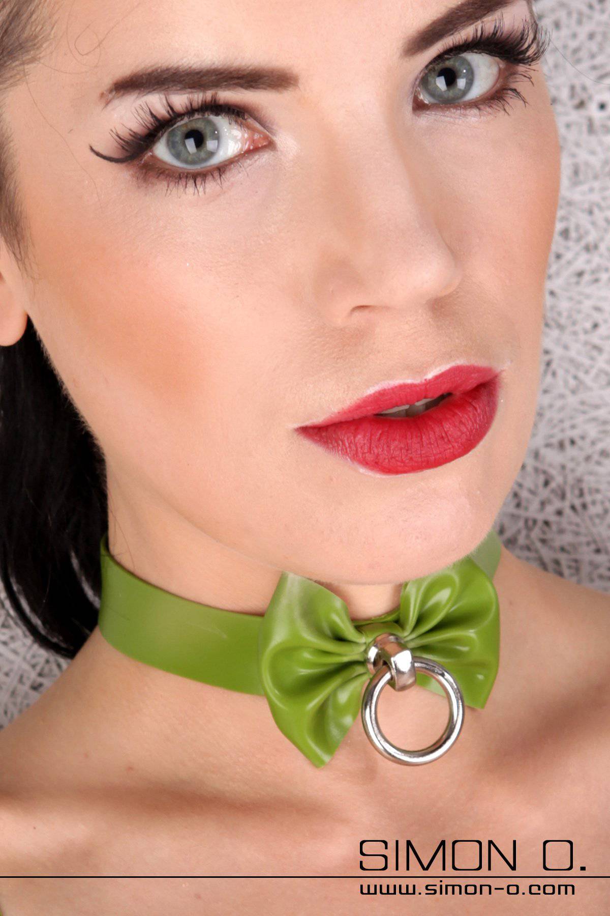 A woman with light make-up wears a green latex collar with bow and O-ring.