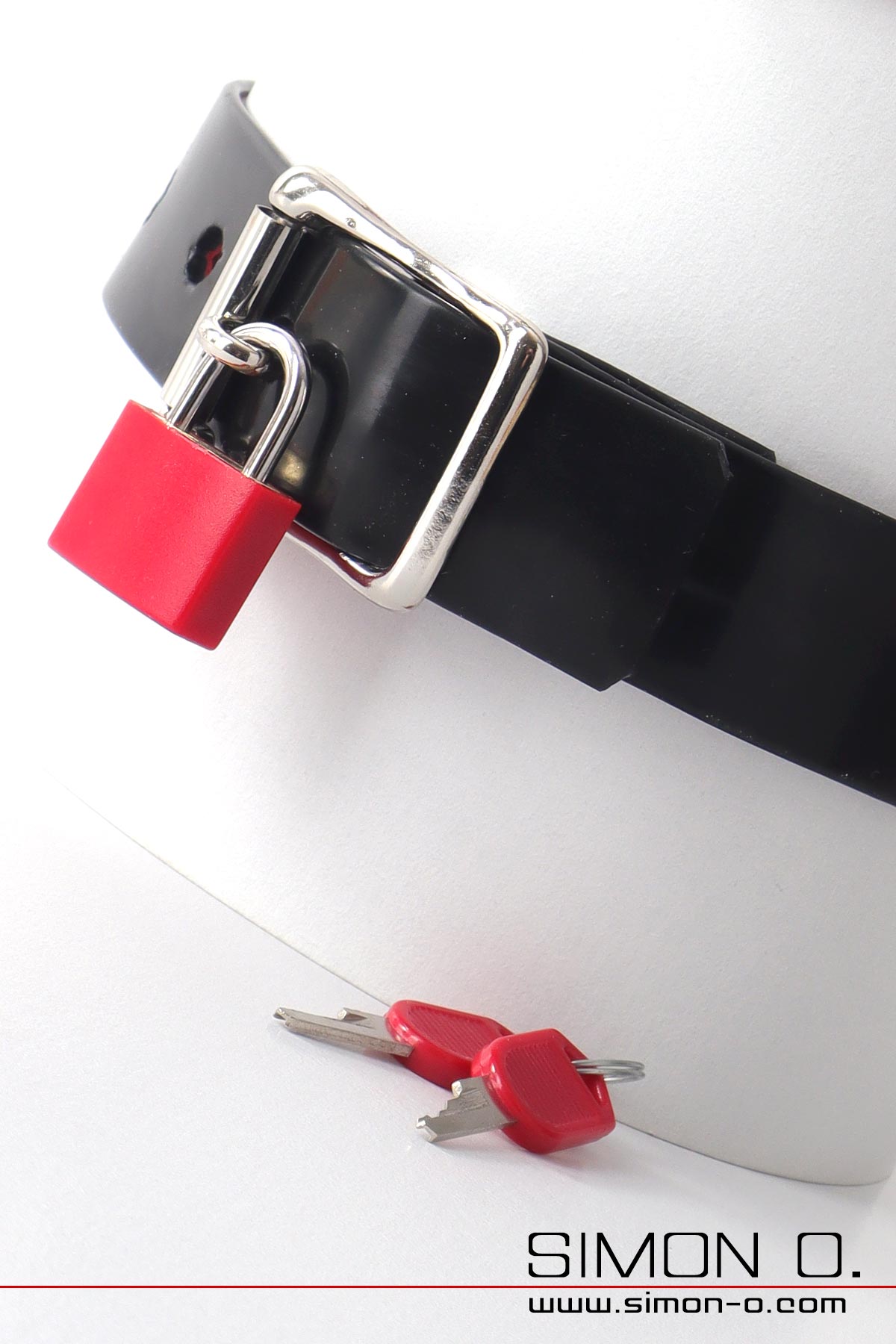 You can see a black latex collar with a silver buckle and red padlock. There are two keys on the white surface underneath.