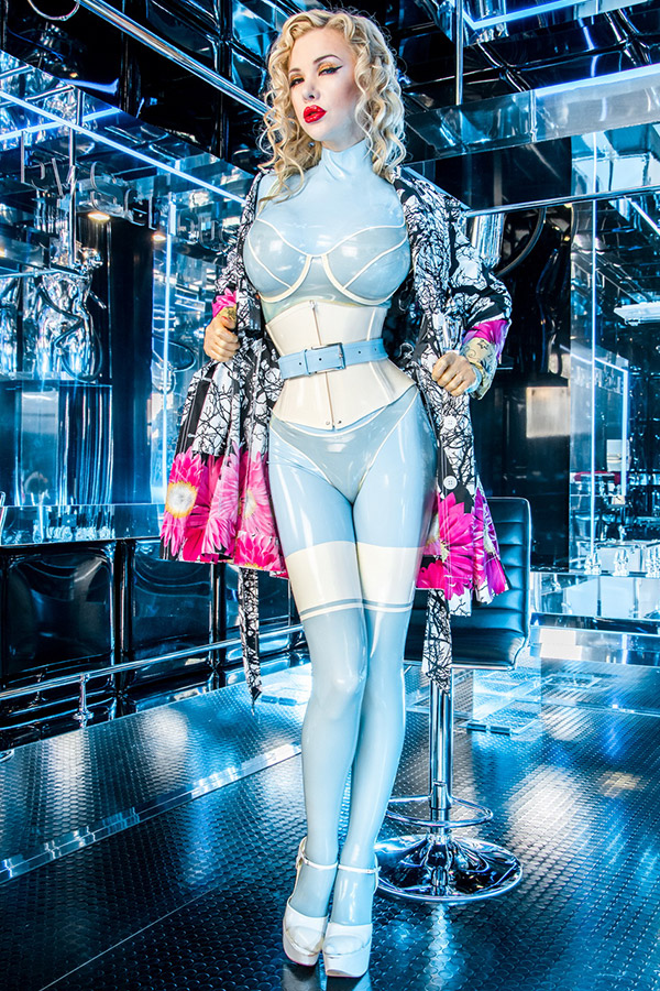 A person in a shiny blue latex catsuit with a corset and high heels stands in a room with shiny surfaces. She holds open a patterned coat with pink accents and reveals latex lingerie which she wears over the catsuit.